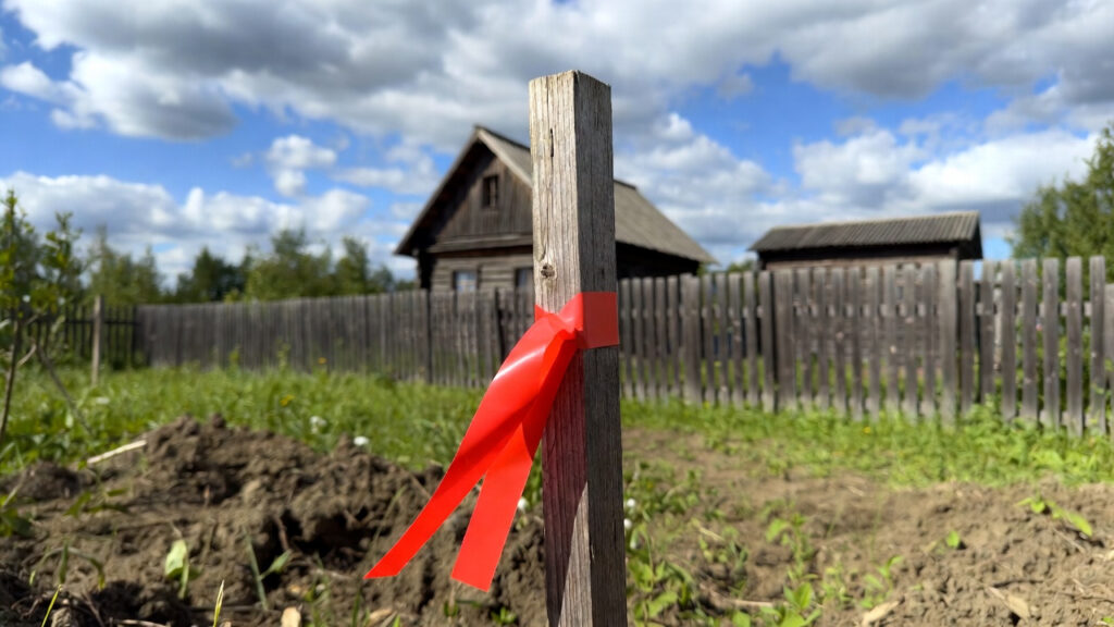 Колышек с красной лентой, обозначающий границу земельного участка на фоне дачного дома.